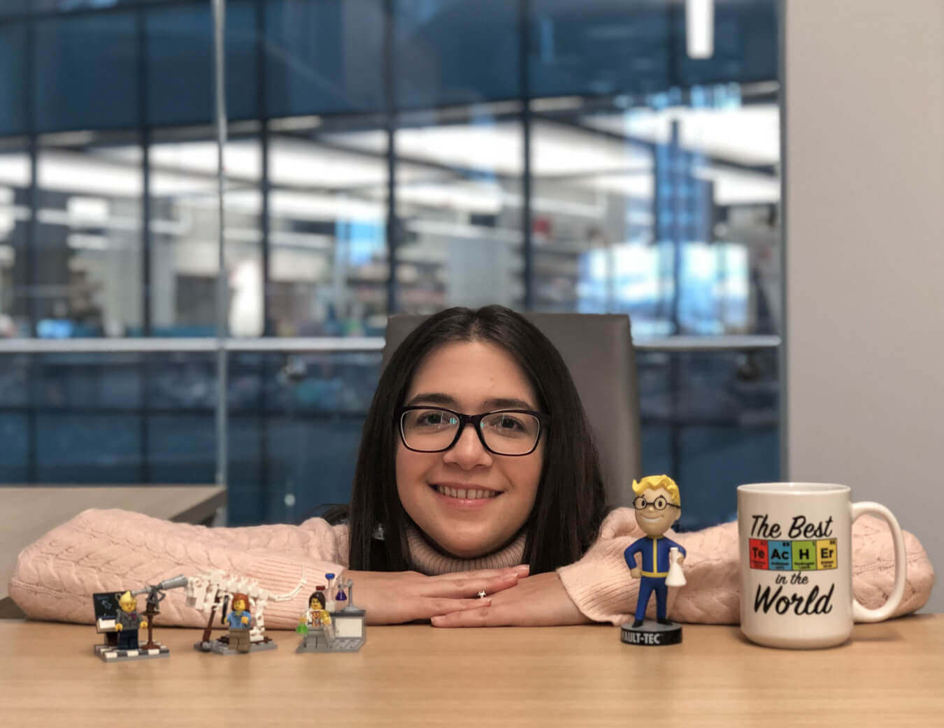 A picture of Amanda Tiano. She is posing for the camera with both arms on a desk and smiling. On the desk with her are various items such as a mug, a vault-boy bobblehead, and lego figurines.