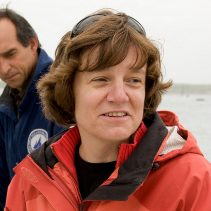 A photo of Dr. Amy Bower posing on camera. Behind her the sky is cloudy and a body of water can be seen. She is wearing a red jacket and has sunglasses on her head.