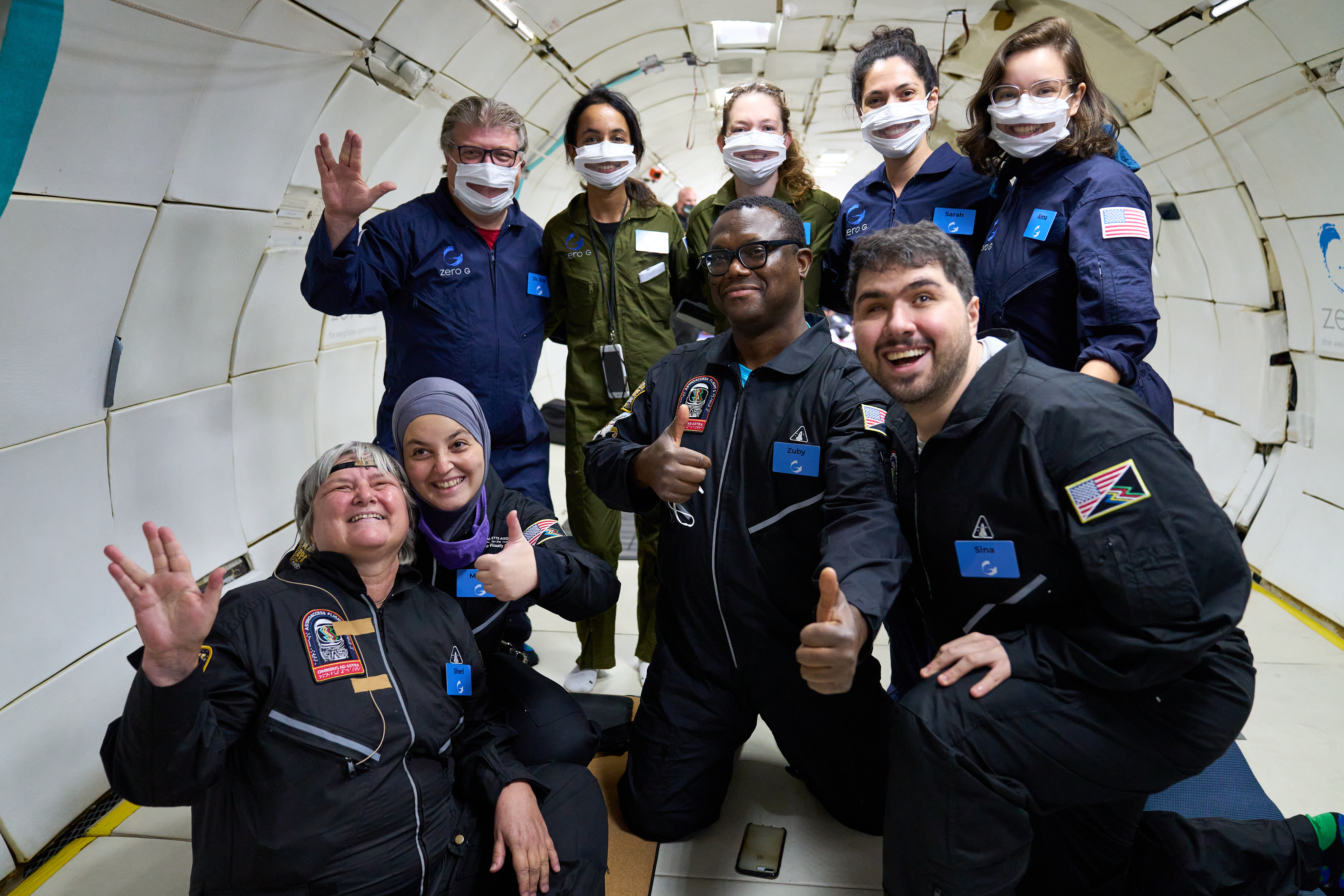Dr. Sheri Wells-Jensen, Mona Minkara, Azubuike Onwuta, and Sina Bahram (left to right) sitting and kneeling on zero gravity plane smiling at camera with 5 support team members also smiling through their cut masks standing behind them. 
                        