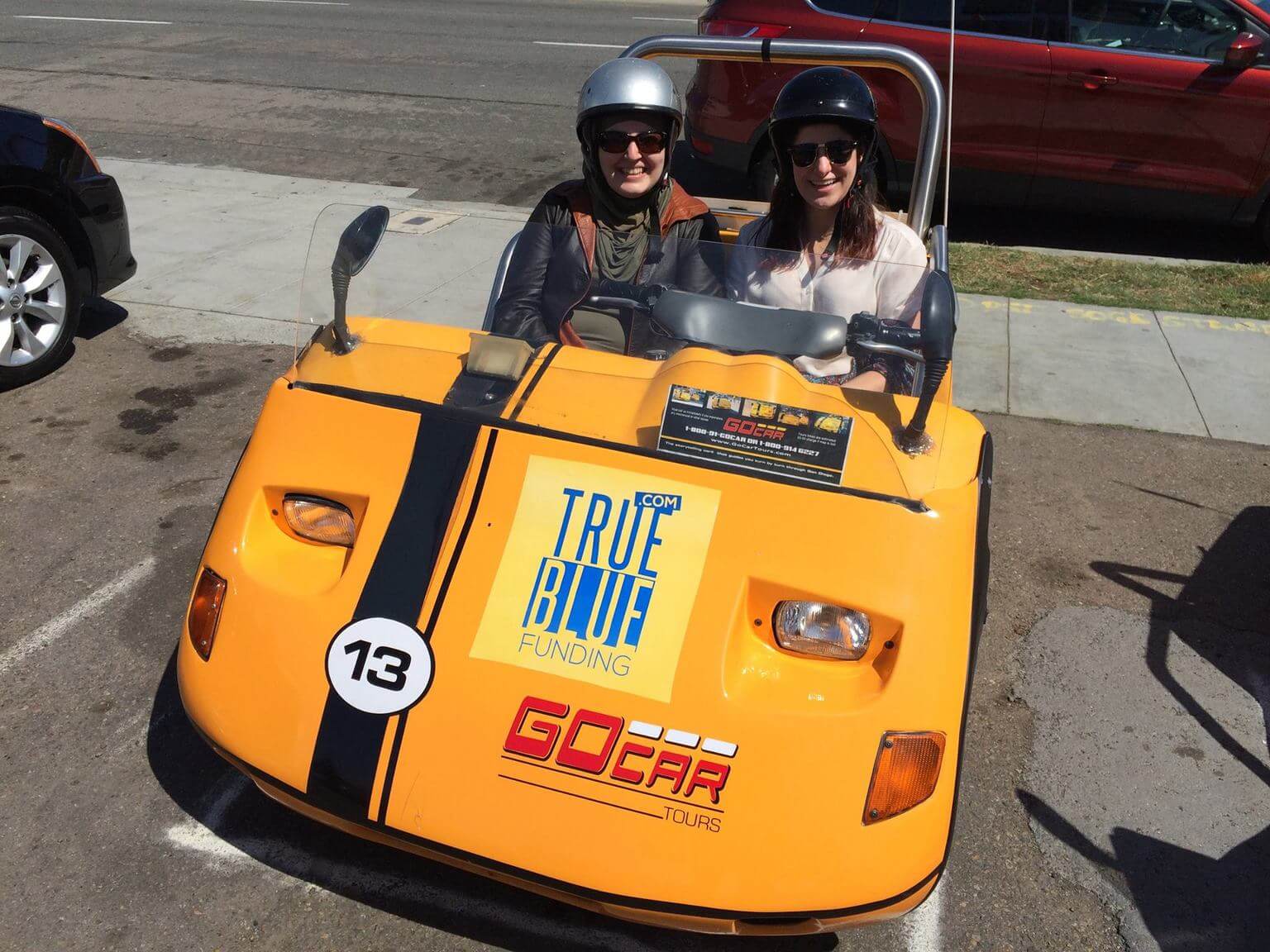 A photo of Mona Minkara and another person in a yellow go kart. Both are wearing helmets and are smiling for the camera. 