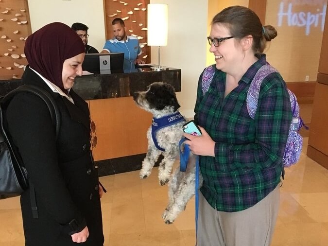 Mona smiling as she meets a small dog that a colleague is holding. They are talking in a hotel lobby.