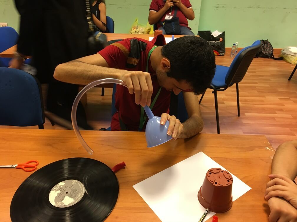  A student with a red shirt sitting at a wooden table. He is looking down as he attempts to attach a clear tube to a funnel. White paper, a small flower pot, and a music record can also be seen on the table. Other students can be seen behind them.