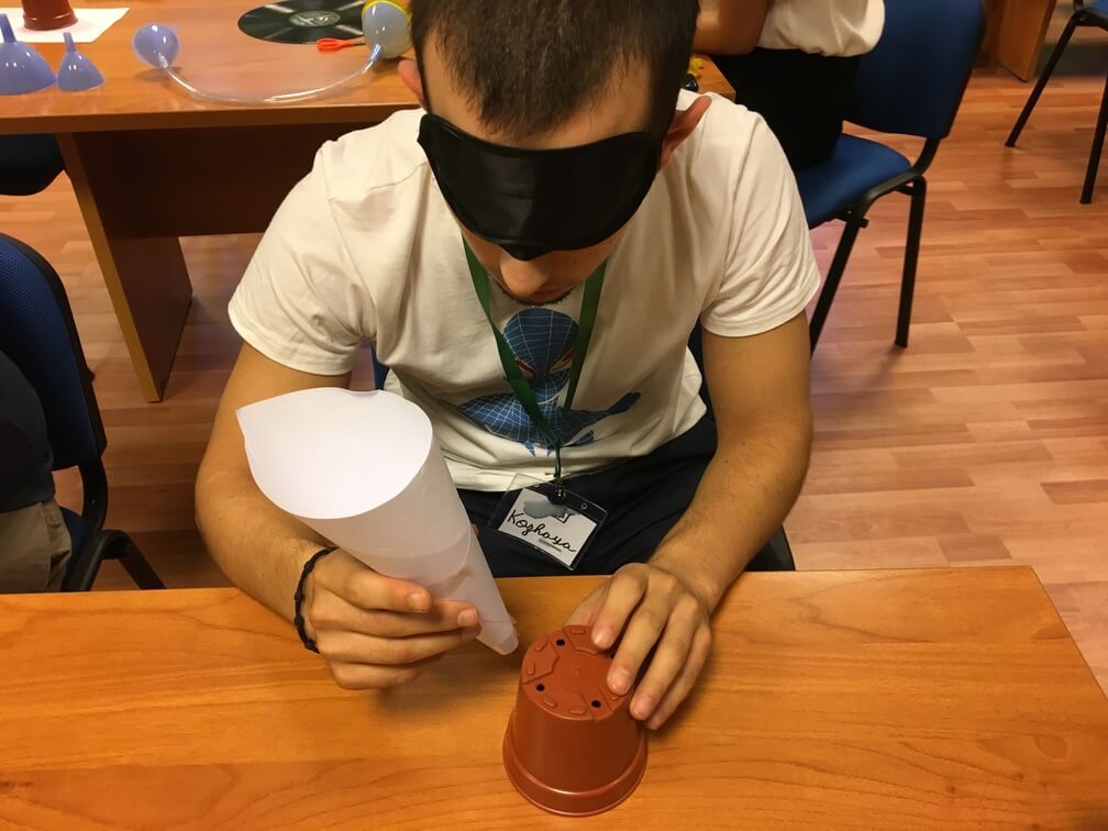 A student wearing a blindfold holds a paper cone in his right hand a flowerpot in the other. He is attempting to attach the cone to the pot. 