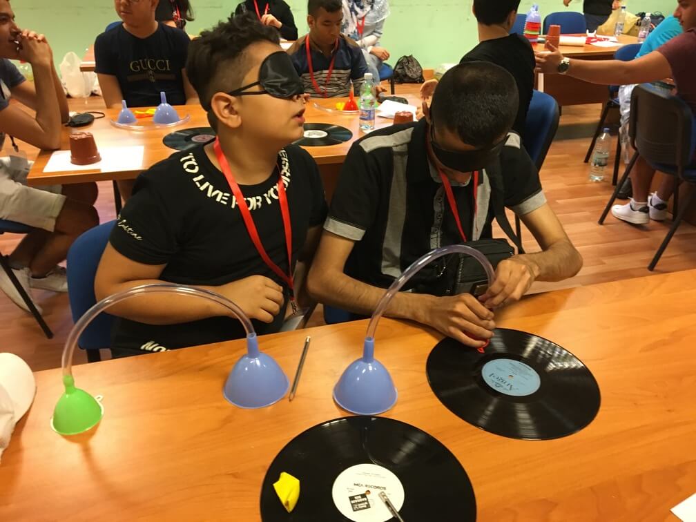 Two students sit at a desk. Both have black blindfolds on. In front of them on the desk are two funnels each connected to a tube. One of the students is currently trying to attach the tube onto a music disk. Other students can be seen behind them.