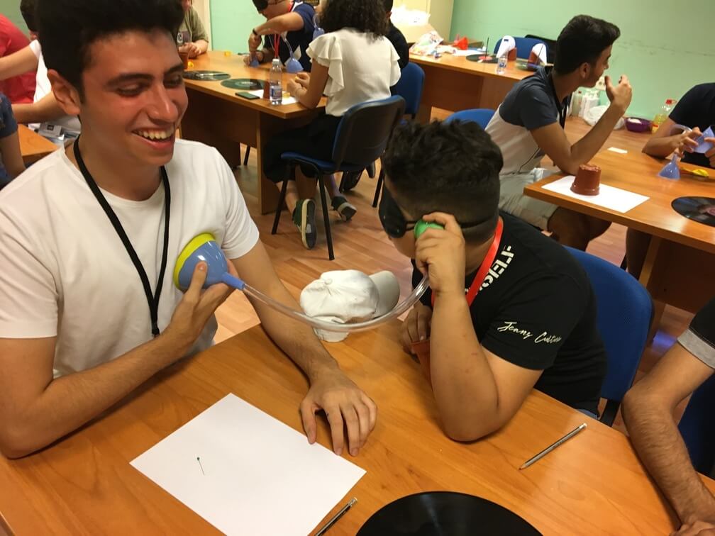A student with a blindfold uses a funnel and tube to listen to the heartbeat of another student.