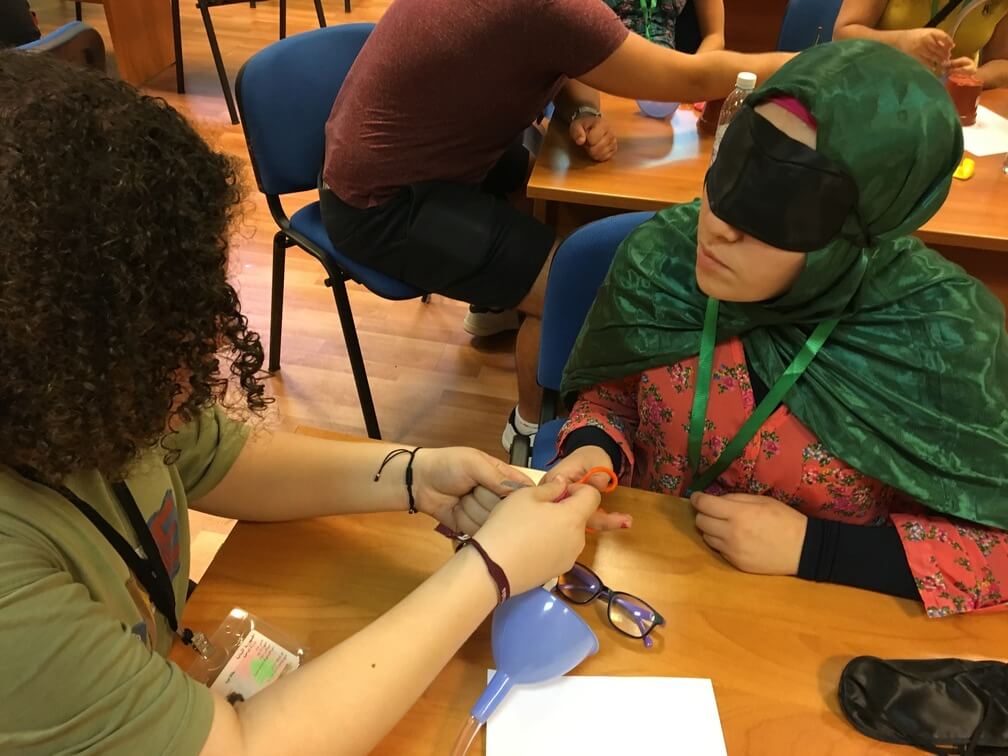 Two students sit at a desk. One wears a green t-shirt while the other wears a pink dress and green hijab. The one in the green t-shirt aid The girl in the pink dress with assembling a contraption as she wears a black blindfold.  Other students can be seen behind them.