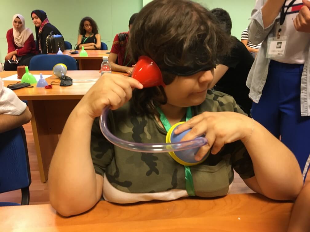 A student wearing a camo T-Shirt uses the tube/funnel contraption to listen to their own heartbeat. With one funnel on their chest and the other up to their ear. Other students can be seen behind them.