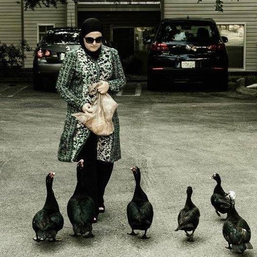 A picture of Mona feeding ducks in a driveway. She is wearing an intricate dress and black slacks. She is holding a bad of food as six black ducks gather around her. 