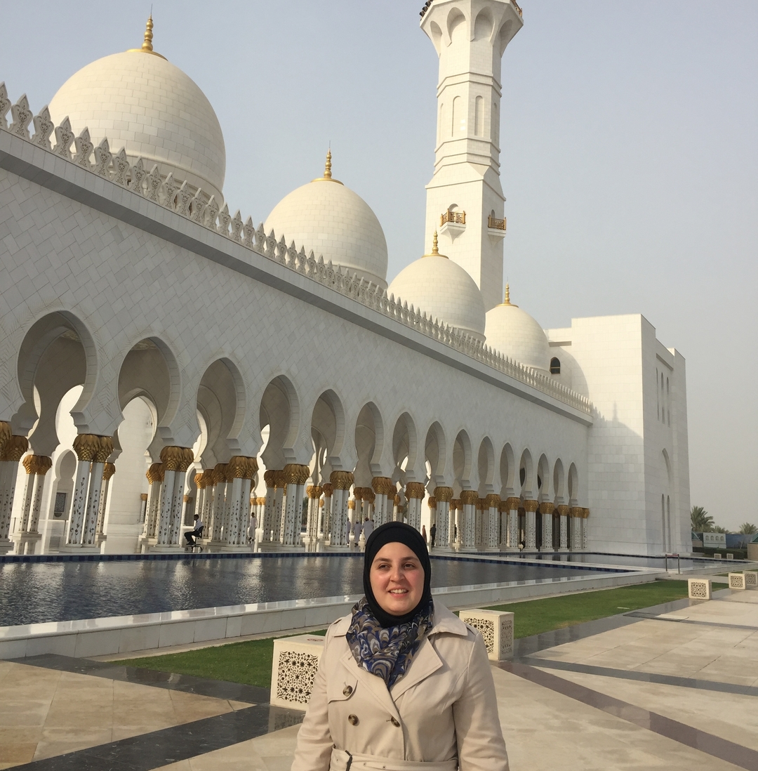 A picture of Mona smiling of the Sheikh Zayed Mosque is Abu Dhabi. She is wearing a beige coat with a black hijab. Behind her the mosque is made of white marble and accented in gold. Next to the mosque is a long reflecting pool. Above the sky is sunny and clear. 