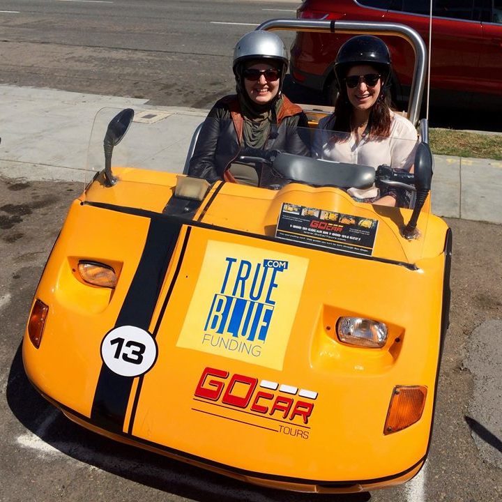 A picture of Mona and a friend in a yellow go-kart. They are both wearing helmets and are smiling for the camera. It is sunny out.