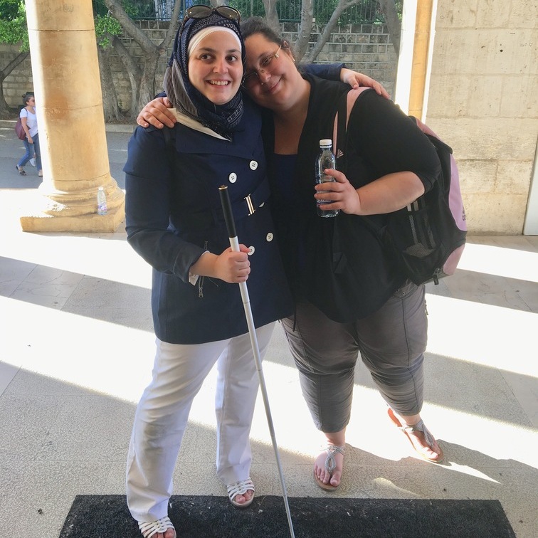 A picture of Mona side-hugging a friend. In one hand she is holding her cane. The friend is holding a water bottle in her free hand. Behind them is a marble pillar and wall. It is sunny and shadows from the marble pillars can bee seen across the floor.