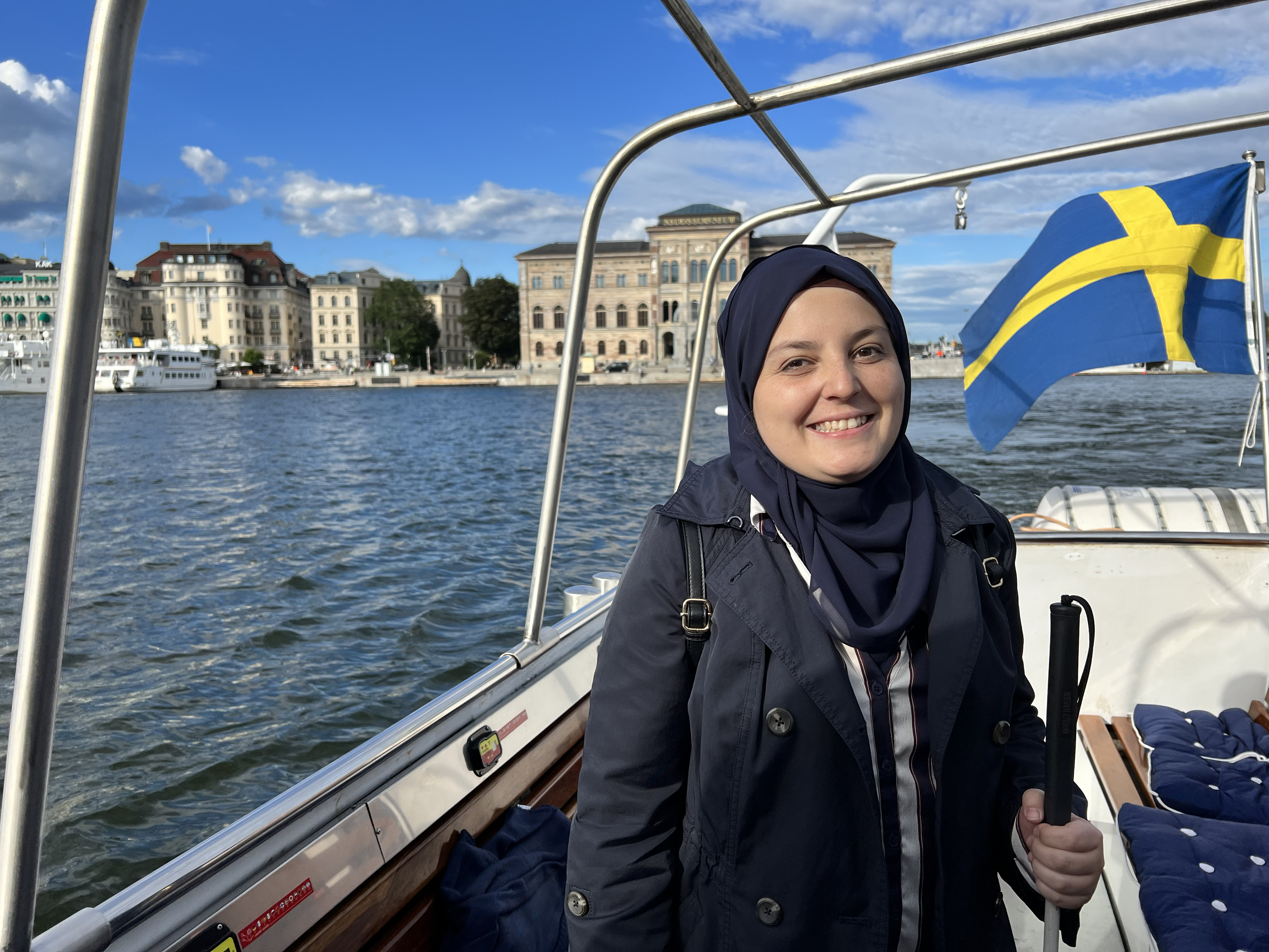A beautiful picture of mona smiling at the camera. She is standing inside a sailing boat facing the camera holding her stick on with her left hand. There is a sweedish flag, attached the boat that is waving in the wind behind her. She is wearing a black coat and a navy blue head scarf. We can see water all around and clear blue skies to the left. We see the harbour at a distance behind her and there are beautiful buildings lined up in the harbour. This looks like a tourist boating location.