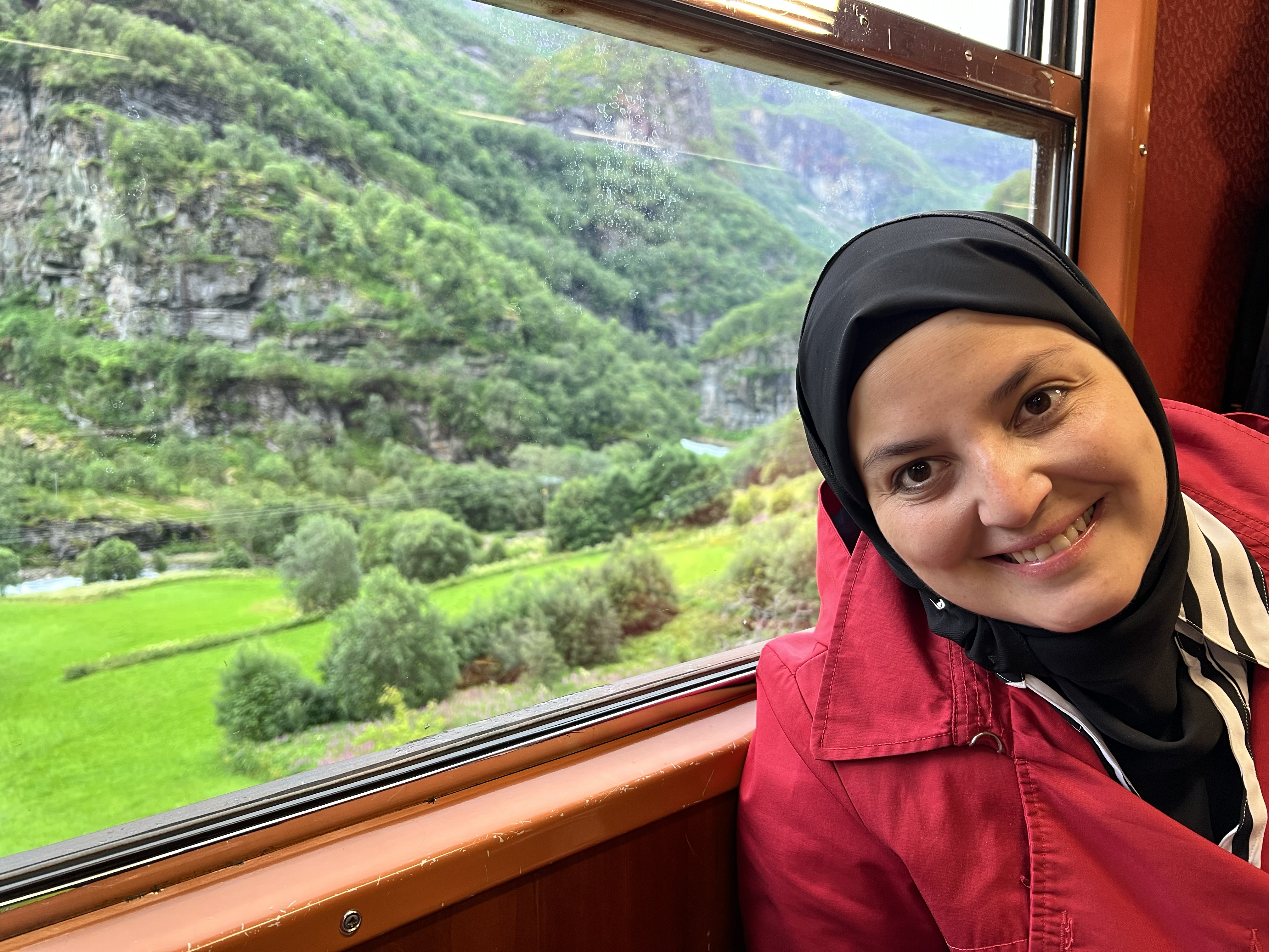 A picture of mona wearing a bright red coat and a black headscarf. She is seated inside a train in her seat and outside the window we are able to see green pastures and hills. She is leaning forward and smiling towards the camera.
