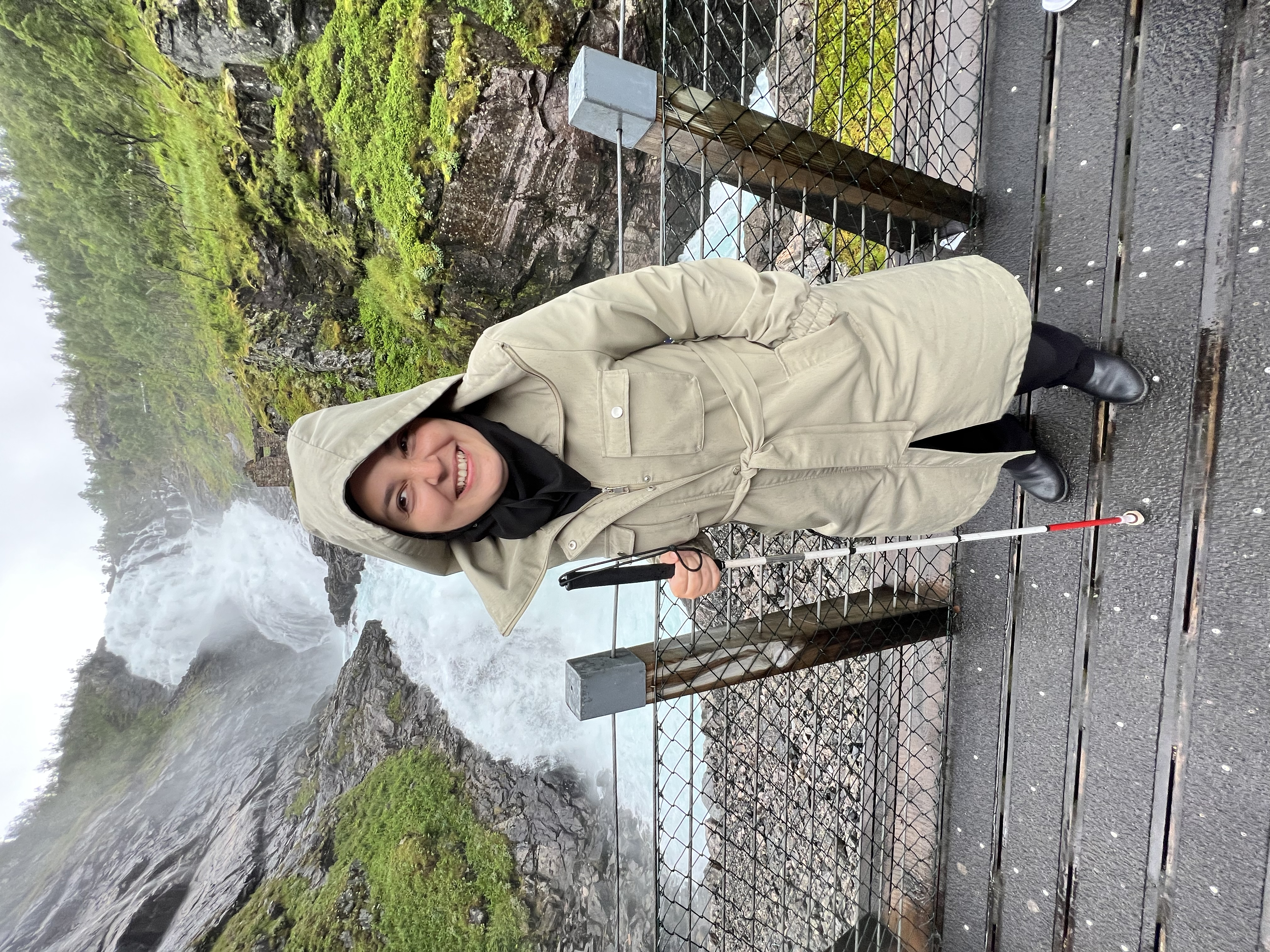 A picture of mona posing for the camera standing on a bridge that is in front of a huge waterfall. We see hills and rocky formations on both sides and the water seems to be falling from a height and flowing underneath the bridge with force. It seems to be pretty cold as mona is wearing a white coat with the hoodie covering her head and her hands are in the coat's pockets. 
