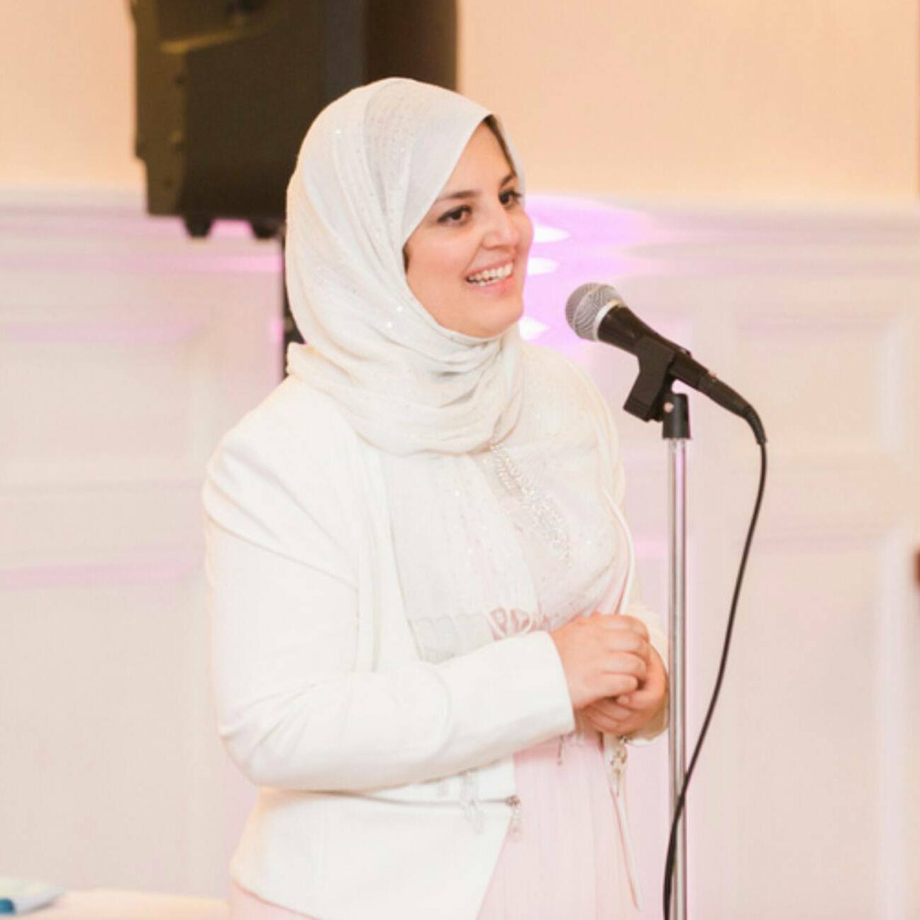 A picture of Mona in an all-white outfit and headscarf. She stands in front of a microphone and is smiling out at an audience.