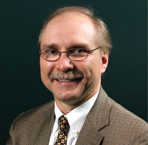 A photo of Dr. Kenneth M. Merz, Jr. smiling for the camera in front of a dark background. He is wearing a brown checkered suit and a white shirt