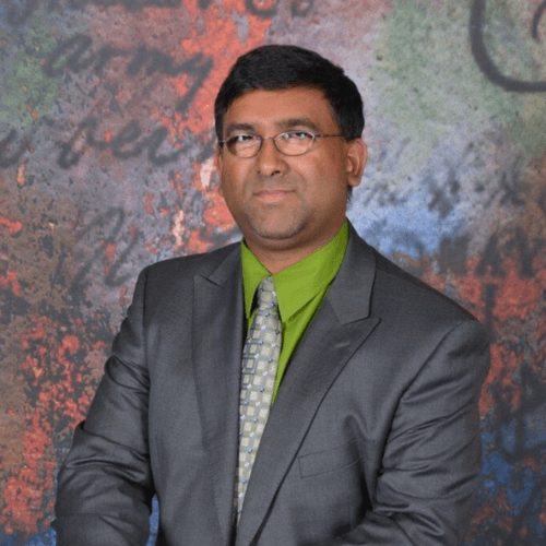 Dr. Mahadeo Sukhai stands on the centre of the image. The image is cut off at his waist. He is indoors and standing in-front of a colourful backdrop, and his hands rest on a white pedestal. He wears glasses, a grey suit, a green shirt, and a light grey tie. His hair is short and dark brown. He smiles softly.