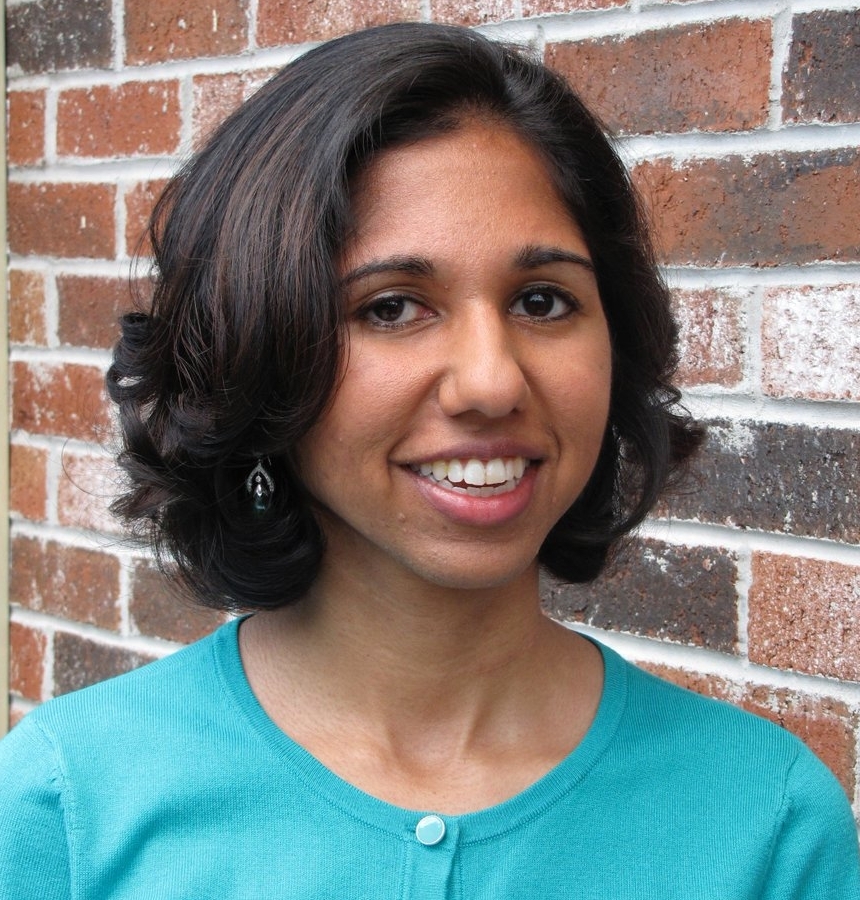 A photo of Dr. Mala Radhakrishnan. She is smiling for the camera and wearing a green shirt. Behind her is a red brick wall.