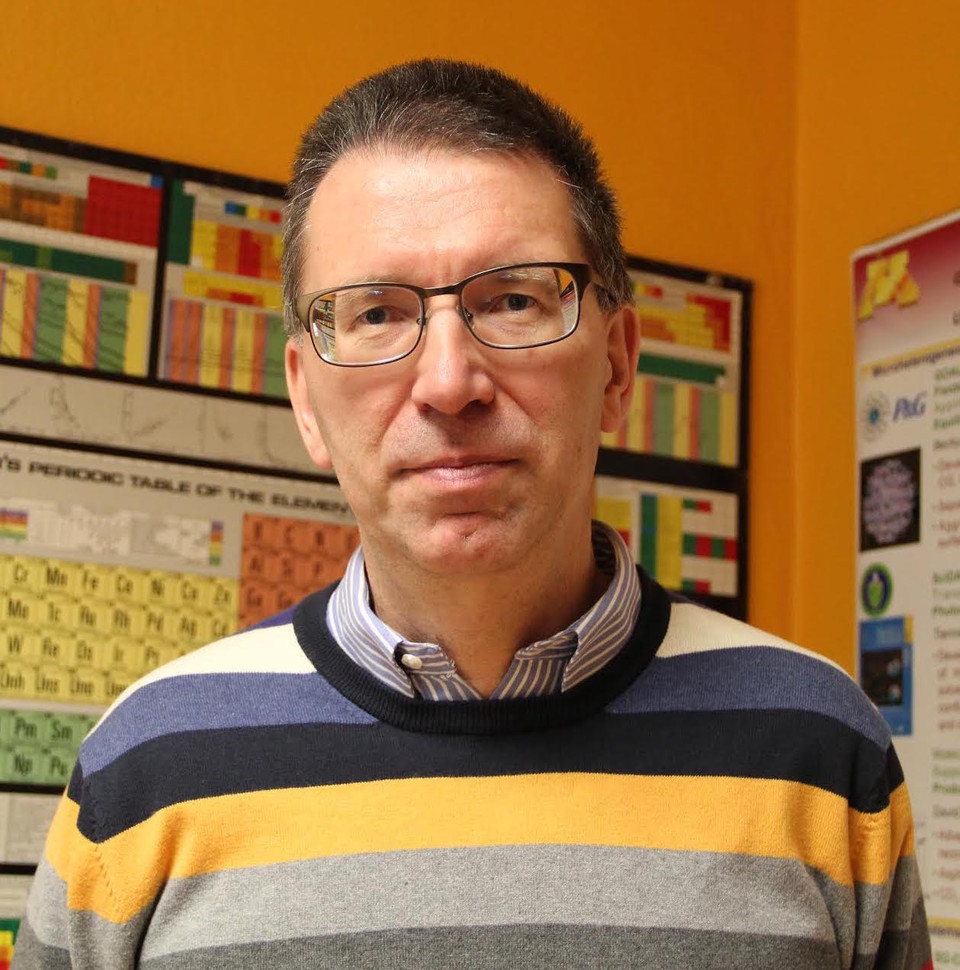 An image of Dr. J. Ilja Siepmann. Dr. Siepmann is wearing a striped sweater over a collared shirt. He wears glasses and is posing for the camera. On the wall behind him is a periodic table and a research poster.