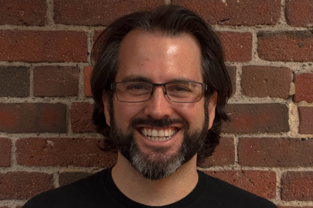 A photo of Kyle Keane smiling brightly in a black tshirt standing against a wall