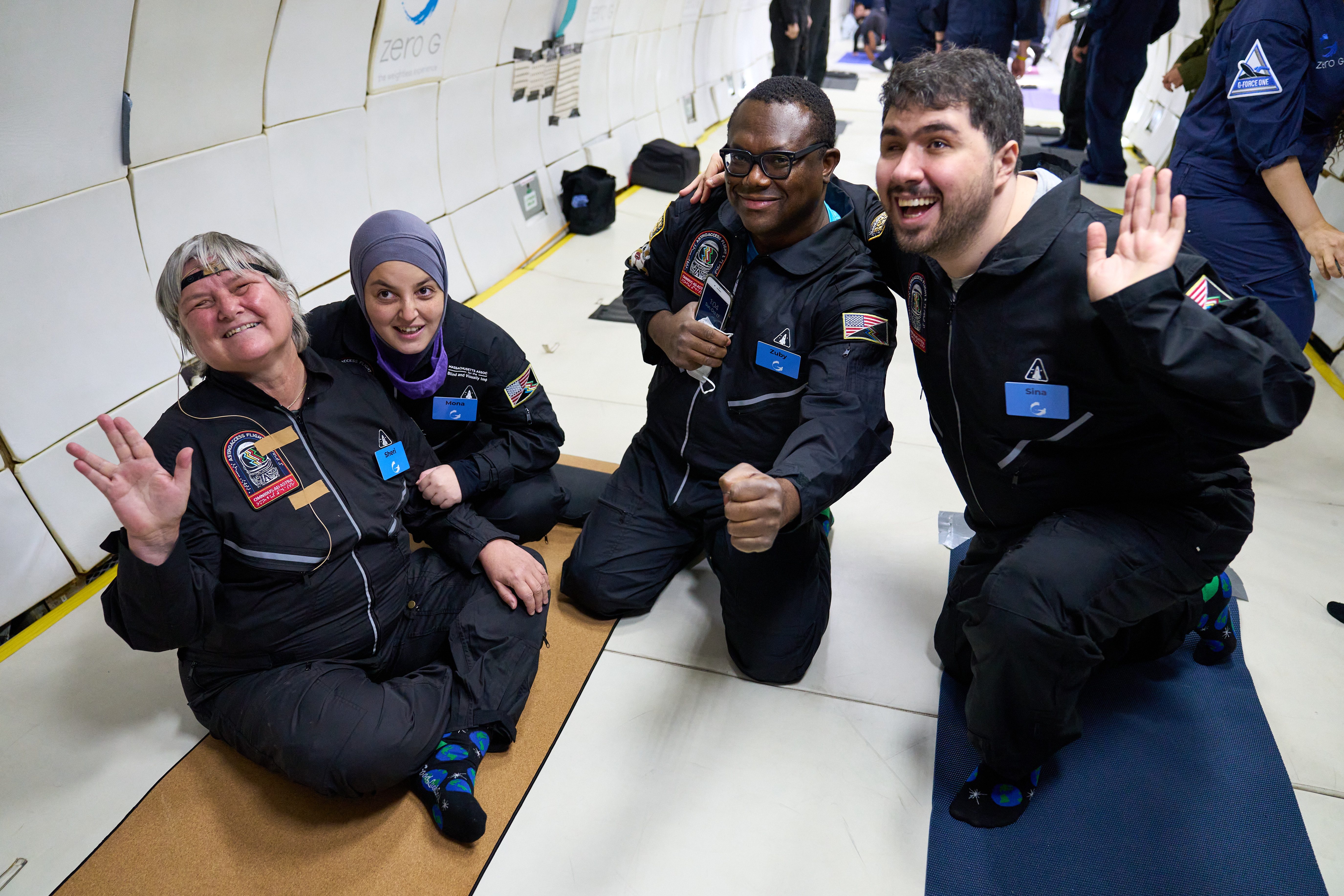 Dr. Sheri Wells-Jensen, Mona Minkara, Azubuike (Zuby) Onwuta, and Sina Bahram (left to right) sitting and kneeling on zero gravity plane smiling at camera.
                            
