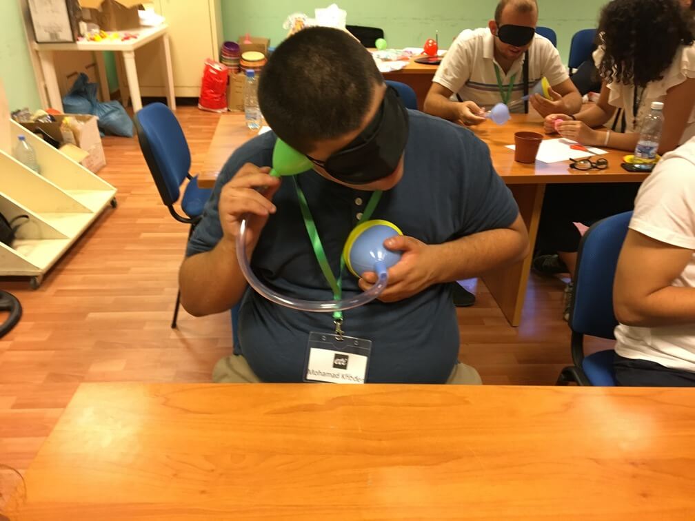 A student wearing a blindfold uses a funnel/tubing contraption to listen to their own heartbeat. Other students can be seen behind him. 