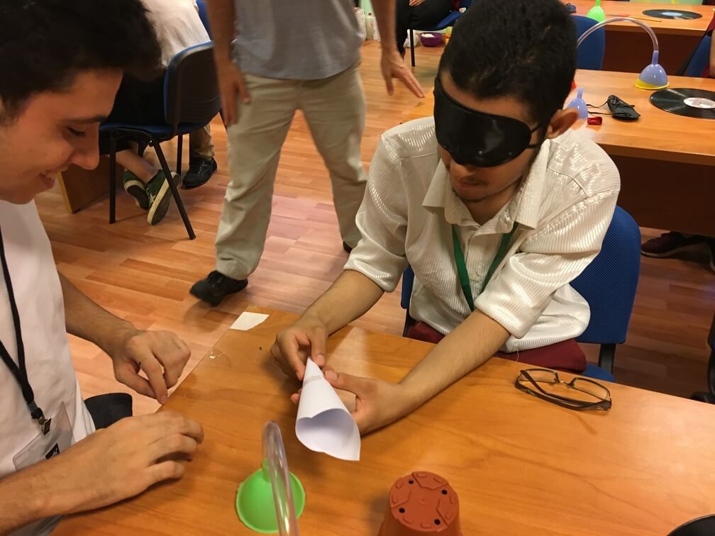Two students attempt to assemble a paper cone. One of them is blindfolded and holding the cone while the other watches him. 