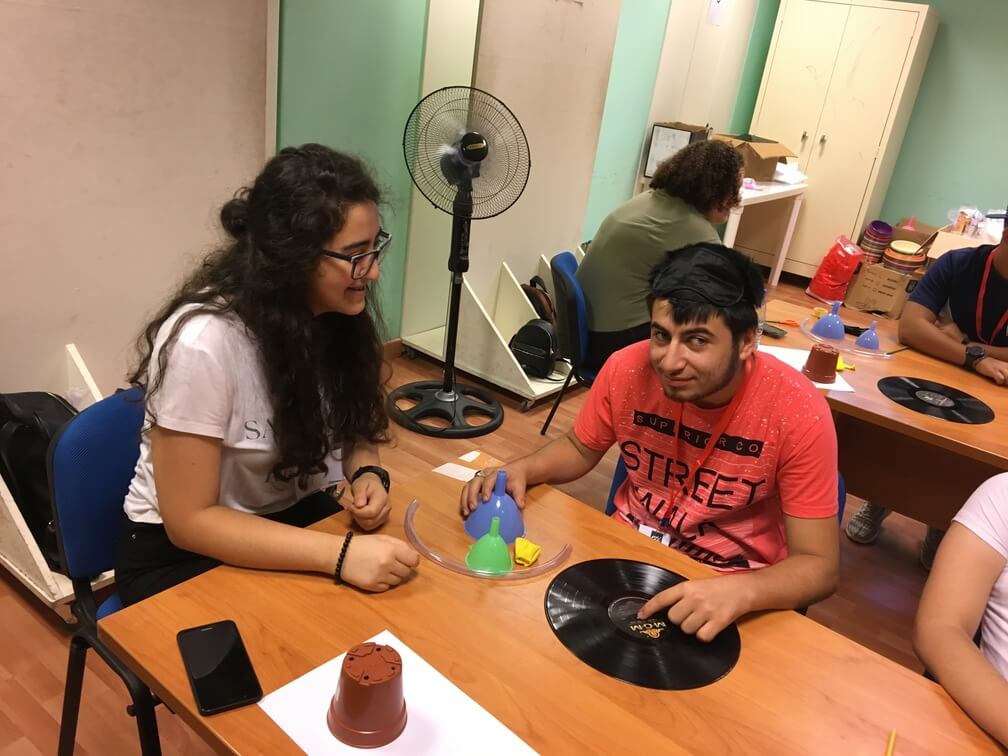 Two students sit at a desk. On the table before them are funnels, tubing, a music record, and a flowerpot. 