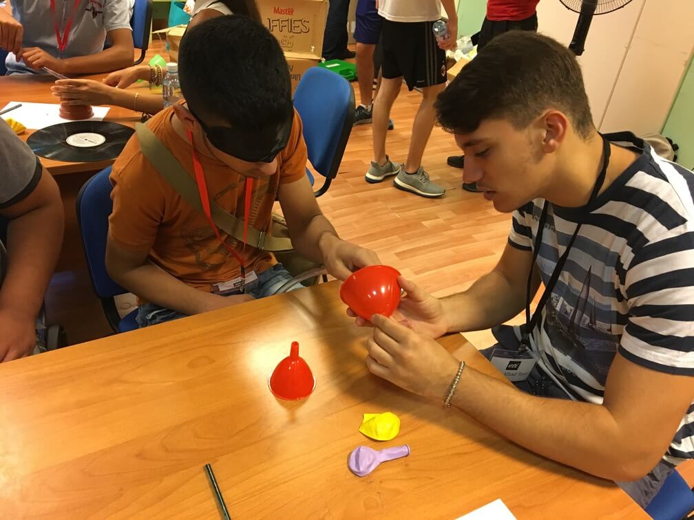 An unblindfolded student aids a blindfolded student with placing a tube into the top of a funnel.