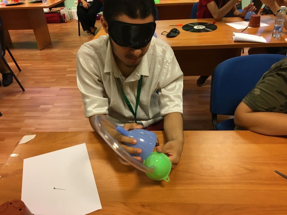 A student in a white shirt wears a black blindfold. He is assembling a contraption that consists of two funnels attached together by clear tubing. Other students can be seen behind them.
