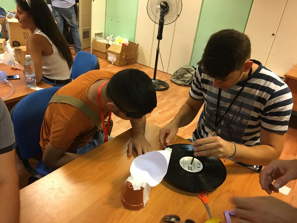 Two students sitting at a desk attempt to assemble a paper music player. One is blind folded while the other puts a paper cone secured by a flowerpot onto a music record. 