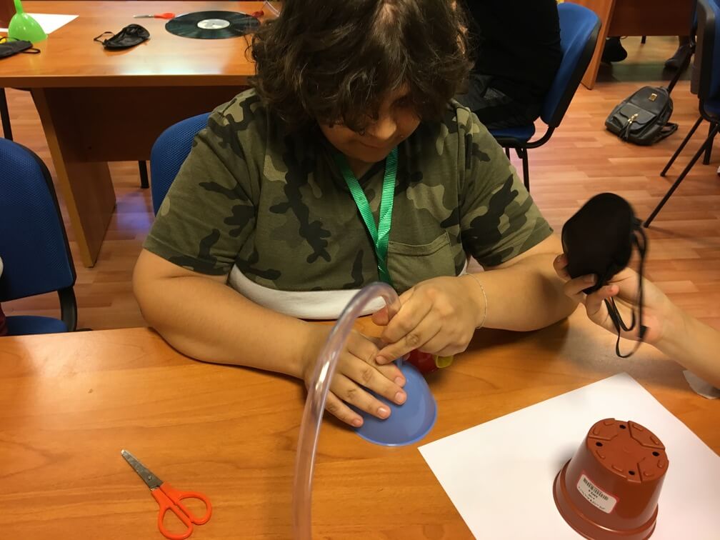 A young student in a camo t-shirt is assembling a clear tube to the bottom of a funnel. A hand to their side is offering them a black blindfold. On the desk there is also a white piece of paper and a small flowerpot. Other students can be seen behind them.