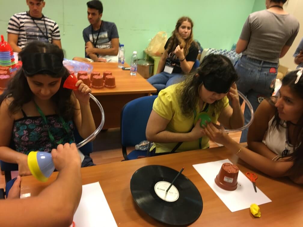 Students sitting at a table. On the right one student wears a blindfold and uses a funnel/tubing contraption to listen to their heartbeat. Another student next to them helps them with this. On the left a student listens into a funnel. On the table between them are music records on a pole.
