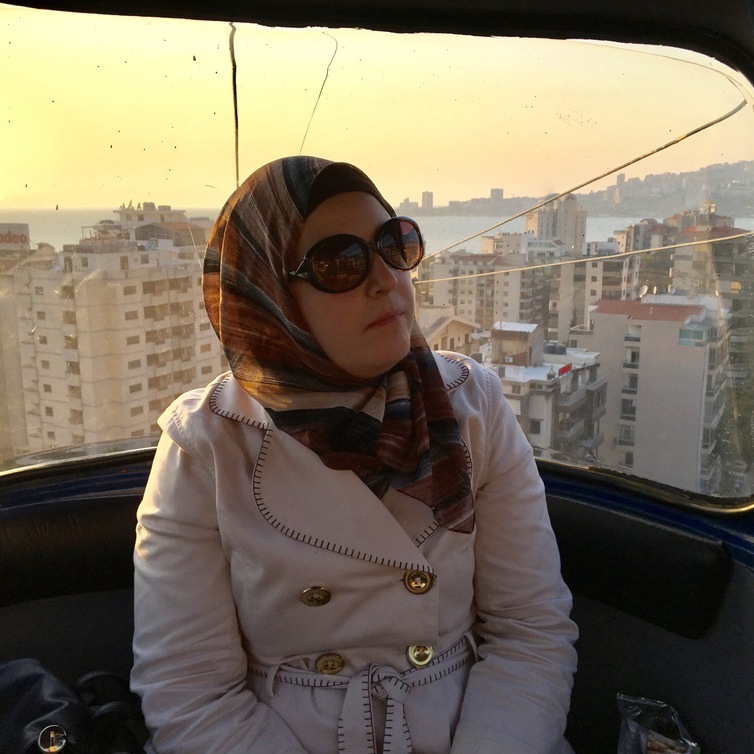 A picture of Mona on a gondola high above a city. She is wearing a white coat and a head scarf. Behind her the sun is setting and tall buildings can be seen. In the distance is a body of water.