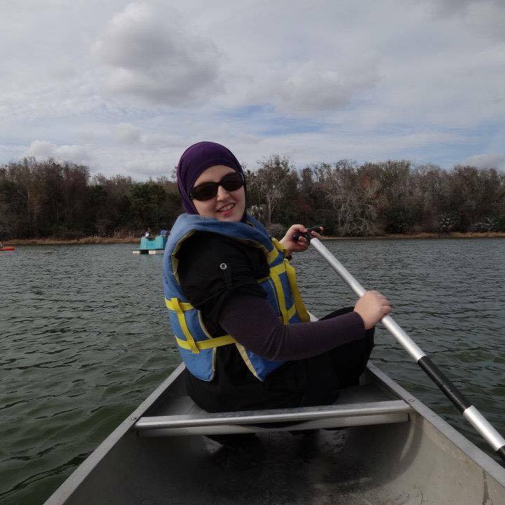 An image of Mona in a small rowboat looking back at the camera smiling. She is wearing a blue life preserver and sunglasses. In her hands is a steel oar. The boat is on a still blue lake during the day.Abve the sky is partially cloudy. 