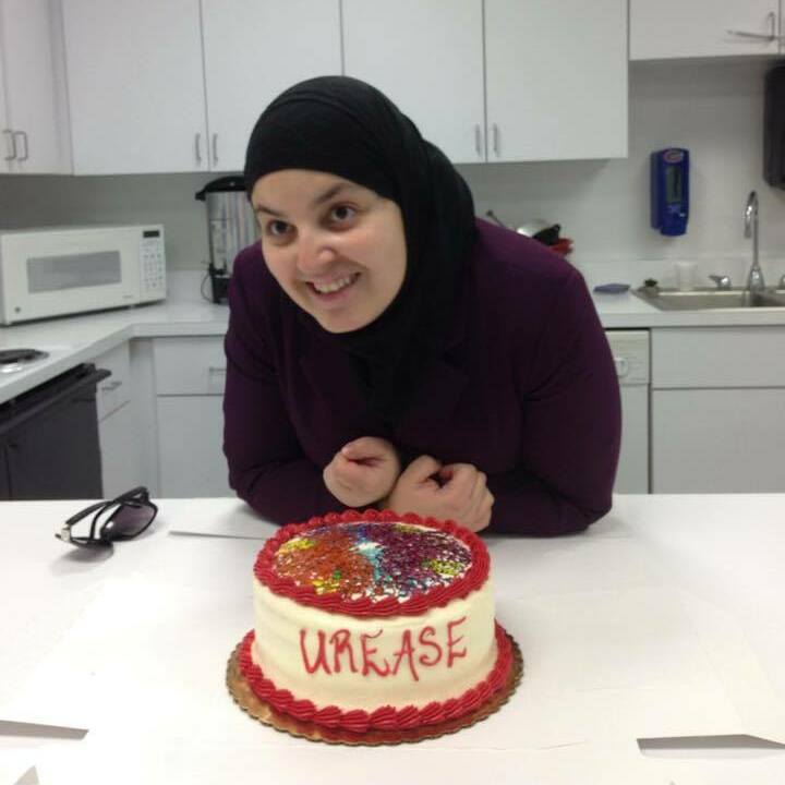 A picture of Mona smiling for the camera. She is wearing a maroon sweater and black hijab, On the counter in front of her is a white and red cake. On top is an intricate design showing the protein structure of Urease in multi-colored frosting. Written in red on the side it reads, “Urease”. Behind her is a plaine white kitchen.
