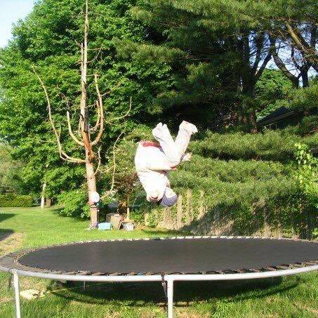A picture of a figure clad in white mid-flip above a trampoline. Behind the figure are green trees. 