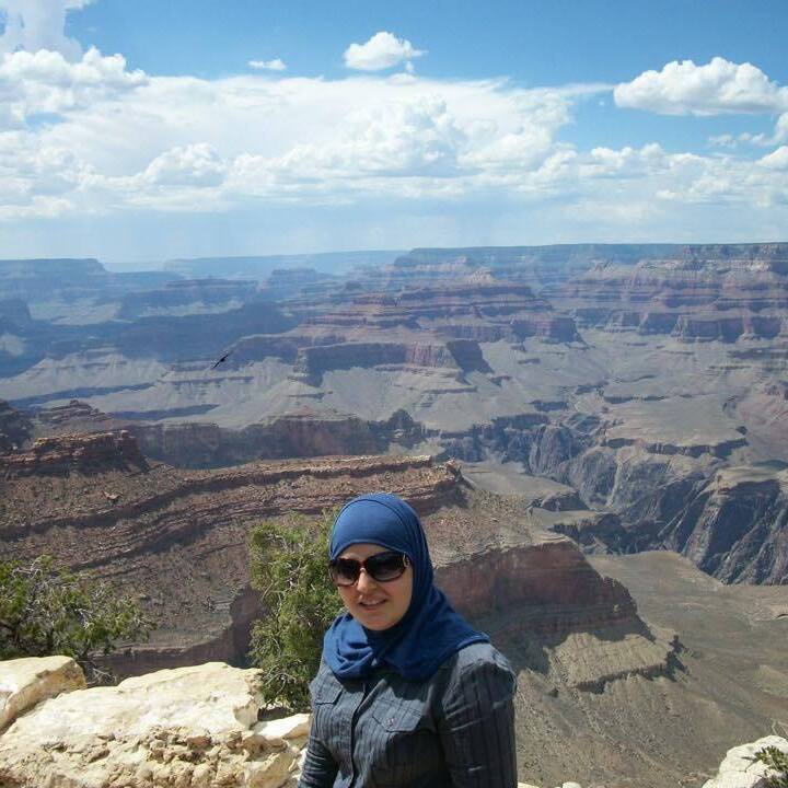 A picture of Mona posing in front of the Grand Canyon. She is wearing a blue dress and hijab. Behind her the canyons stretch for miles. And above the sky is a blue with large white clouds. 