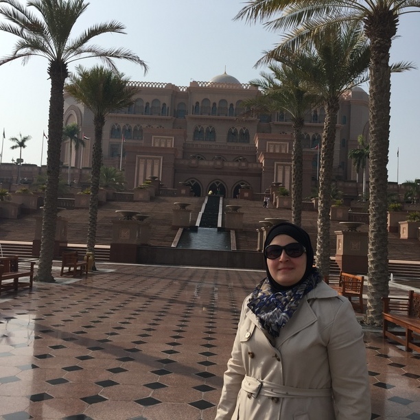 A picture of Mona posing in front of the Emirates Palace hotel in Abu Dhabi. She is wearing a beige coat with a black hijab. On either side of her are tall palm trees. The path to the hotel is paved with a checkered pattern of marble title. Above the sky is clear. 