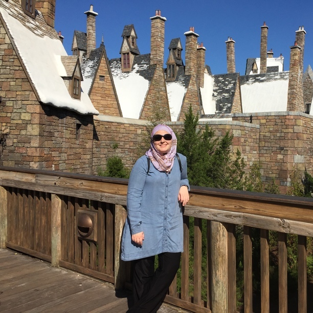 An image of Mona posing for the camera on a wooden bridge. She is wearing a blue shirt and black slacks with a pink head scarf. Behind her is a row of buildings built of brick with large chimneys coming out the top. Each roof has fake snow on top. Above the sky is a deep clear blue and it is sunny.