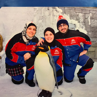 An image of mona and two others posing behind of a penguin. All three are wearing a red, blue, and black jumpsuit. All around them the surrounding is white with snow. All three wear either a headscarf or a hat.