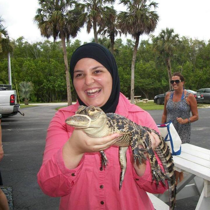 A picture of mona in a pink shirt and black hijab. She is smiling and in her hands is a small baby Alligator. Behind her are palm trees and the sky is cloudy. 