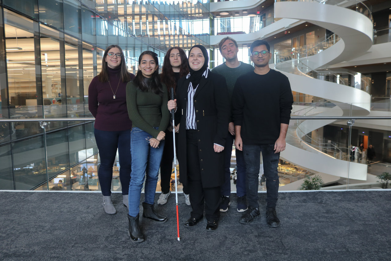 A picture of the Mink Inc. team. From left to right is Amanda Tiano, Anjali Chandani, Anxhela Becolli, Mona MInkara, Benjamin Roy Greenvall, and Afridi Shaik. They are standing in front of a large open atrium with glass windows and a twisting staircase.