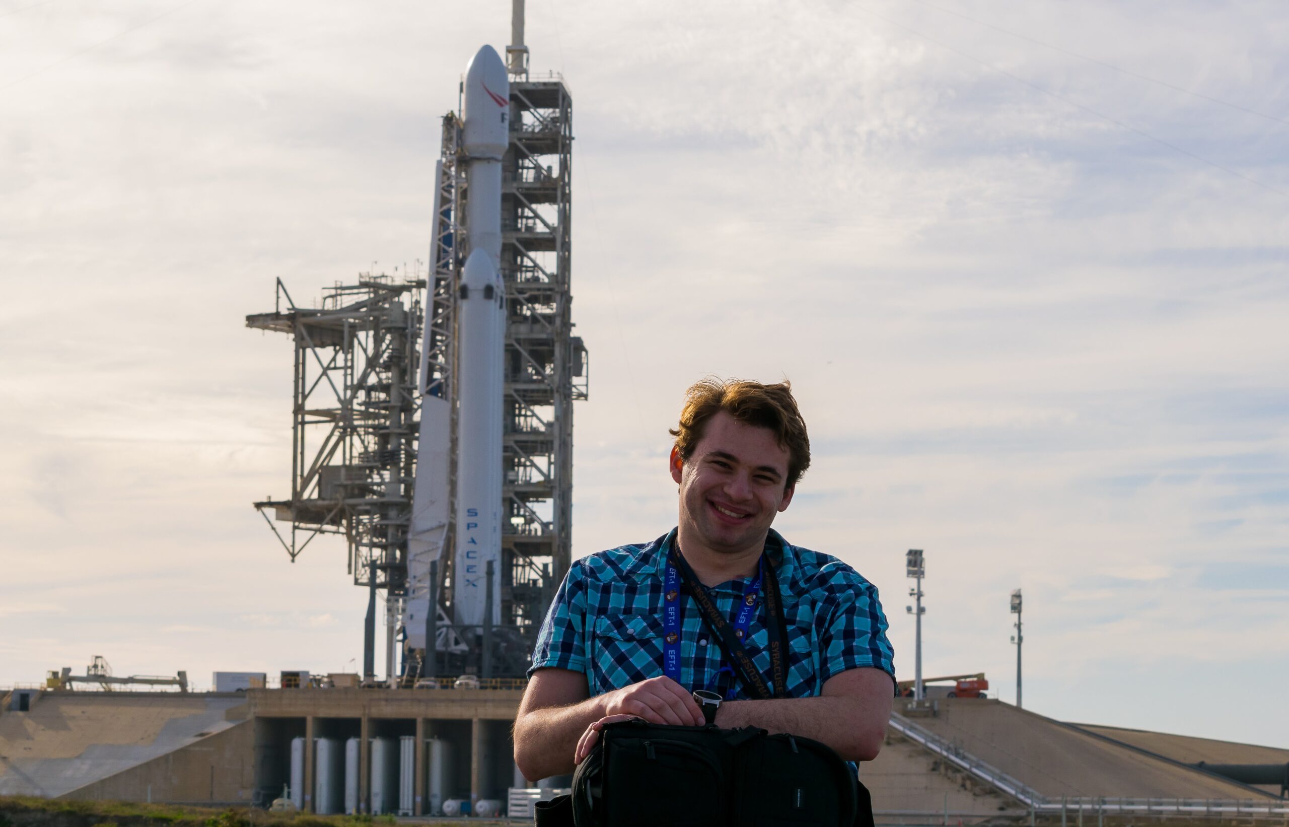 A photo of Sawyer Rosenstein positioned off-center slightly to the right smiling at the camera with his hands on top of a bag. On left behind him there is a SpaceX rocket.