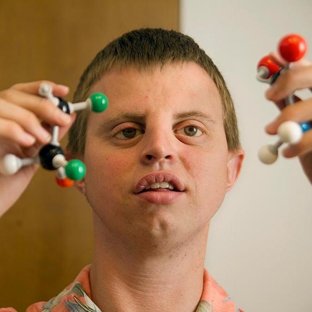 A photo of Dr. Henry Wedler. He is holding a ball and stick model of a molecule in each hand. Behind him the background Is brown and white.